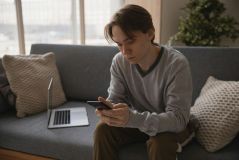 a boy sitting next to a laptop using his phone
